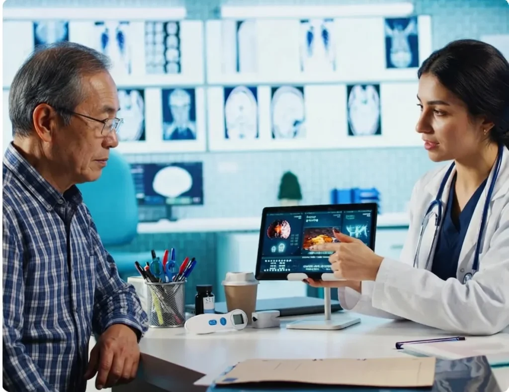 An elderly man in a plaid shirt listening to a female doctor in a white coat who is pointing at a tablet displaying medical scans.